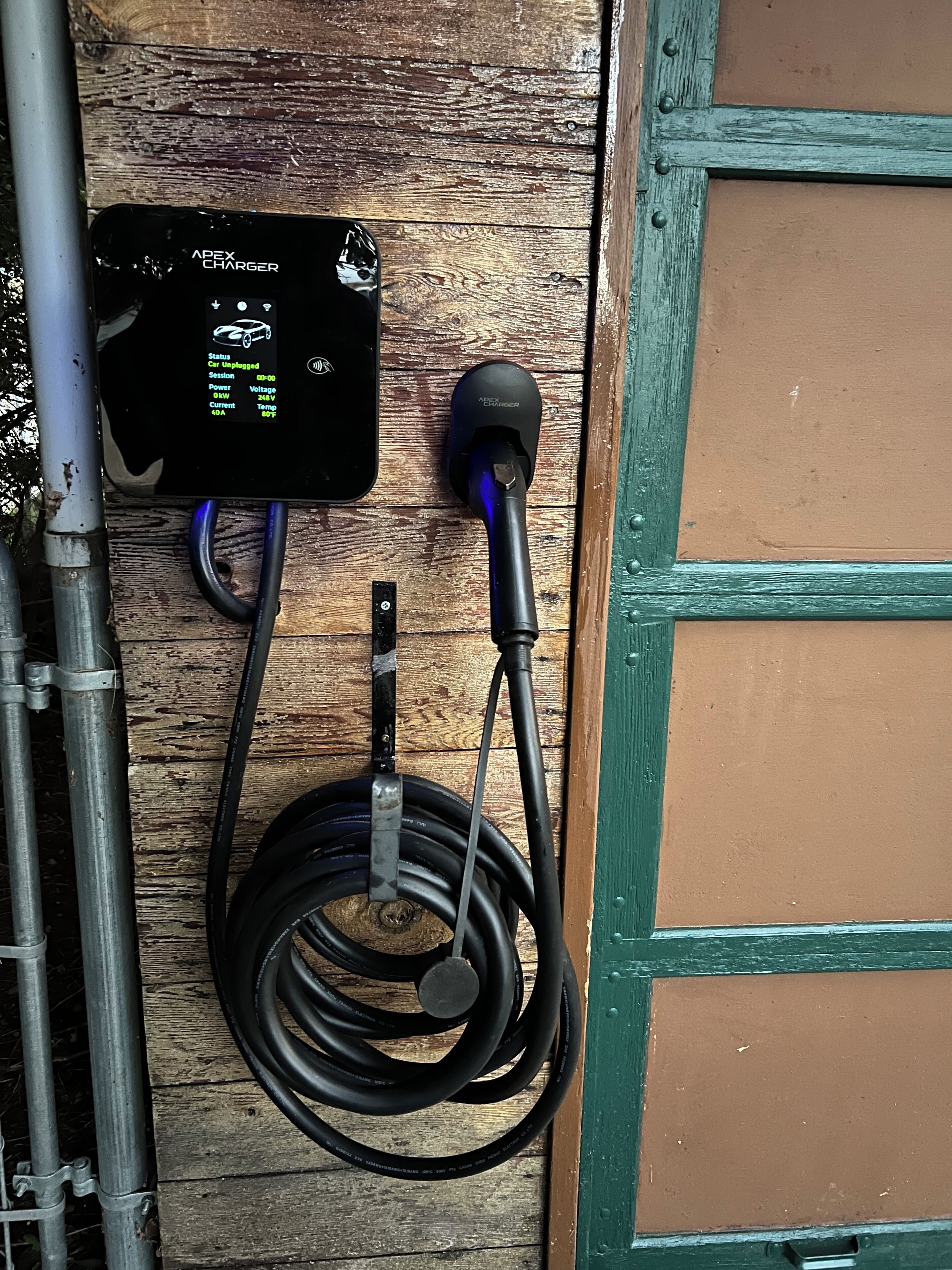 An Apex Charger device mounted on a wooden wall with a coiled charging cable.