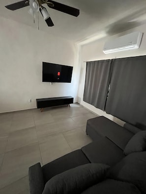 Una sala de estar moderna con un televisor de pantalla plana montado en la pared, un ventilador de techo, un ventanal con cortinas y un sofá de color oscuro.