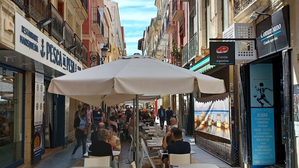 Ein Straßencafé mit Tischen und Stühlen unter einem großen Sonnenschirm, umgeben von Gebäuden mit Balkonen und Beschilderung.