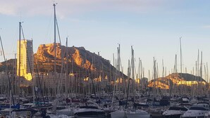 Ein Yachthafen mit zahlreichen vertäuten Booten, ein markantes Gebäude im Hintergrund und eine gebirgige Landschaft.