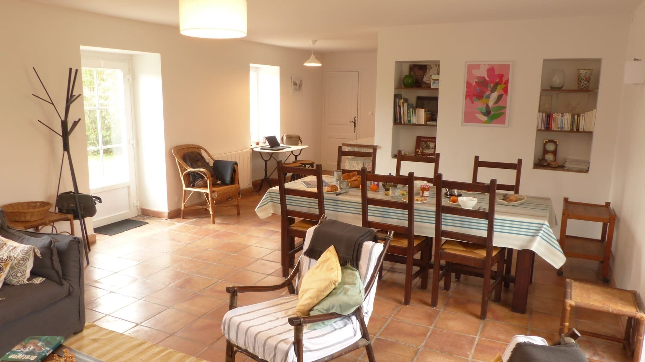Un área de comedor con una mesa preparada para cuatro personas, rodeada de sillas de madera. Se observa un sofá y una silla con cojines al frente.