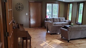 A spacious living room with wooden flooring, a piano, a large clock, and a sofa set.