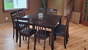 A dining area with a wooden table and chairs, a window with a view of a house and trees, and a decorative mirror on the wall.