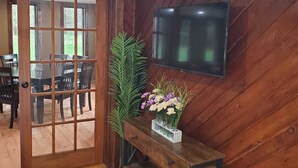 A room with a wooden floor, a TV mounted on the wall, a wooden bench with a vase of flowers, and a door leading to another room.