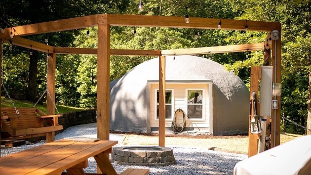 A wooden pergola with hanging lights, a stone fire pit, and a covered outdoor seating area with a picnic table.