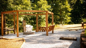 A wooden pergola with seating, surrounded by gravel and trees.