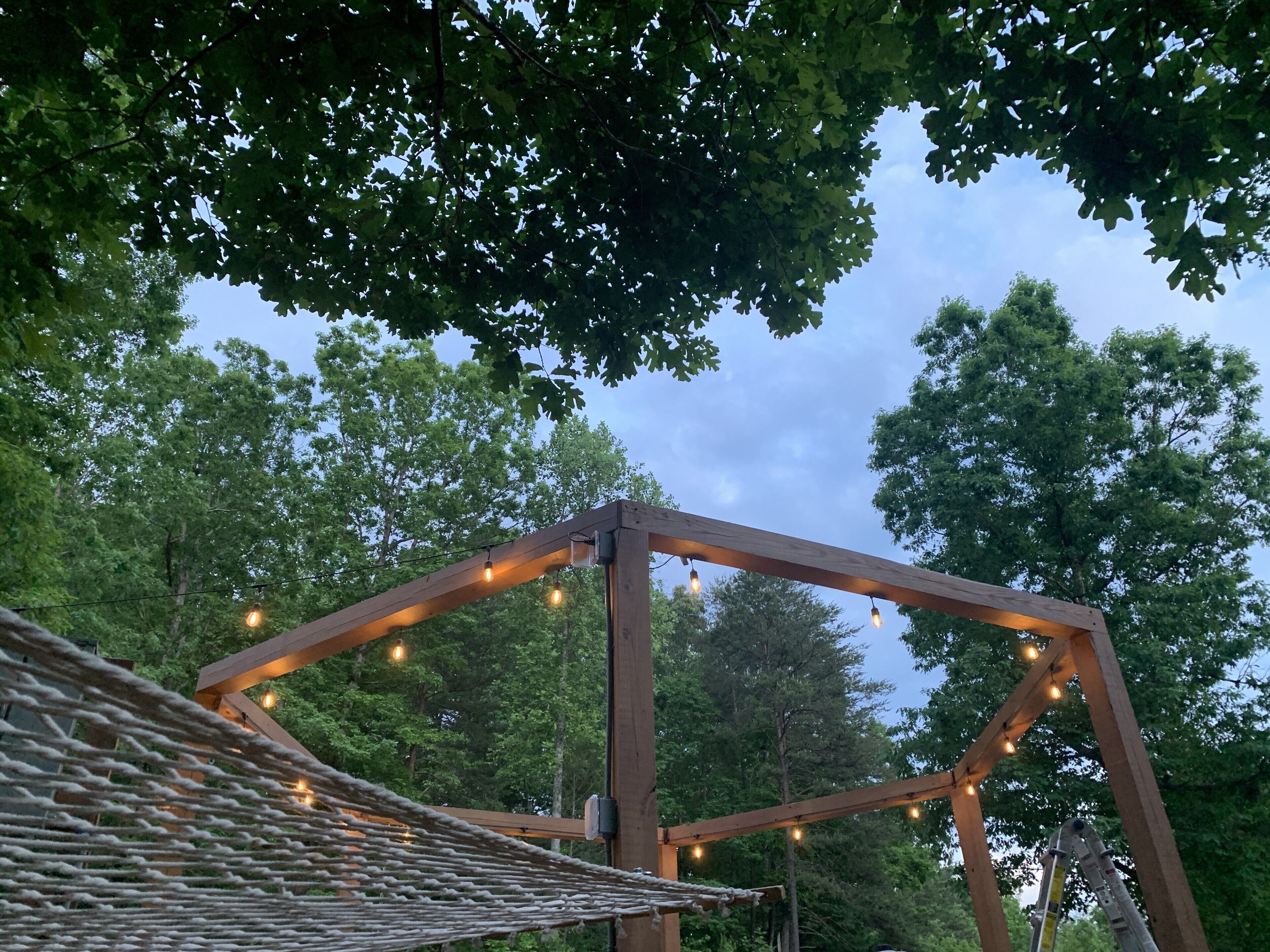 A wooden pergola structure with string lights, surrounded by trees.