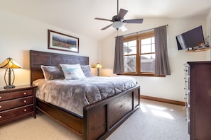 A bedroom with a wooden bed, two bedside tables with lamps, a ceiling fan, a TV mounted on a wooden shelf, and a window with curtains.