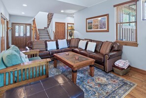A spacious living room with a brown leather sectional sofa, a wooden coffee table, and a patterned rug. A staircase is visible in the background leading to another level.