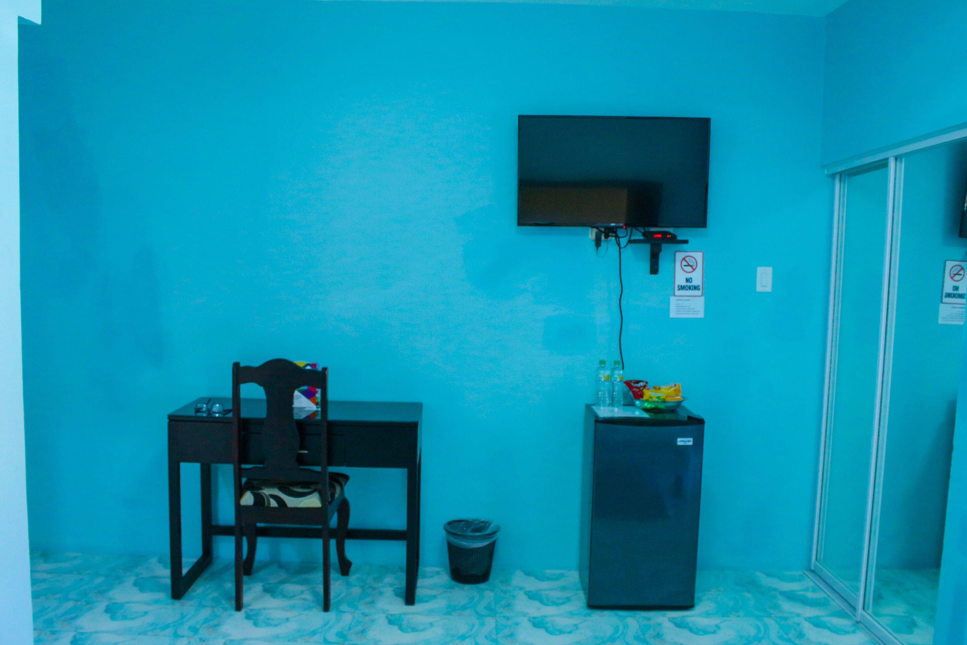 Une petite pièce avec un mur bleu, un bureau avec une chaise, un téléviseur fixé au mur, une poubelle et un réfrigérateur.