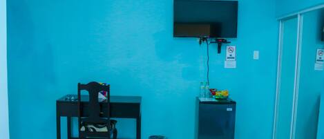 Une petite pièce avec un mur bleu, un bureau avec une chaise, un téléviseur fixé au mur, une poubelle et un réfrigérateur.