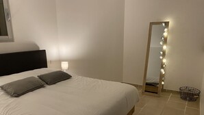A neatly made bed with white bedding and two pillows, a bedside lamp, a full-length mirror with lights, and a small black basket on the floor. - Lovely 2-room apartment in the heart of Esterel with swimming pool (Les Adrets-de-l'Estérel)