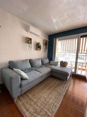A living room with a grey sectional sofa, a woven rug, and wall-mounted shelves with plants.