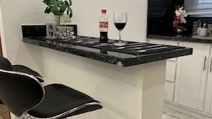 Une cuisine moderne avec un plan de travail noir, des armoires blanches et un sol en marbre. On y trouve deux tabourets de bar noirs et une plante en pot sur le plan de travail.