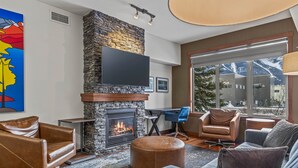 A modern living room with a stone fireplace, leather armchairs, a grey sofa, and a large window offering a mountain view.