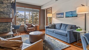 A modern living room with a stone accent wall, a large window with a mountain view, a blue sofa, leather chairs, and a small table with a lamp.