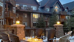 A cozy outdoor seating area with Adirondack chairs and a fire pit, surrounded by a stone fireplace and a snow-covered ground.