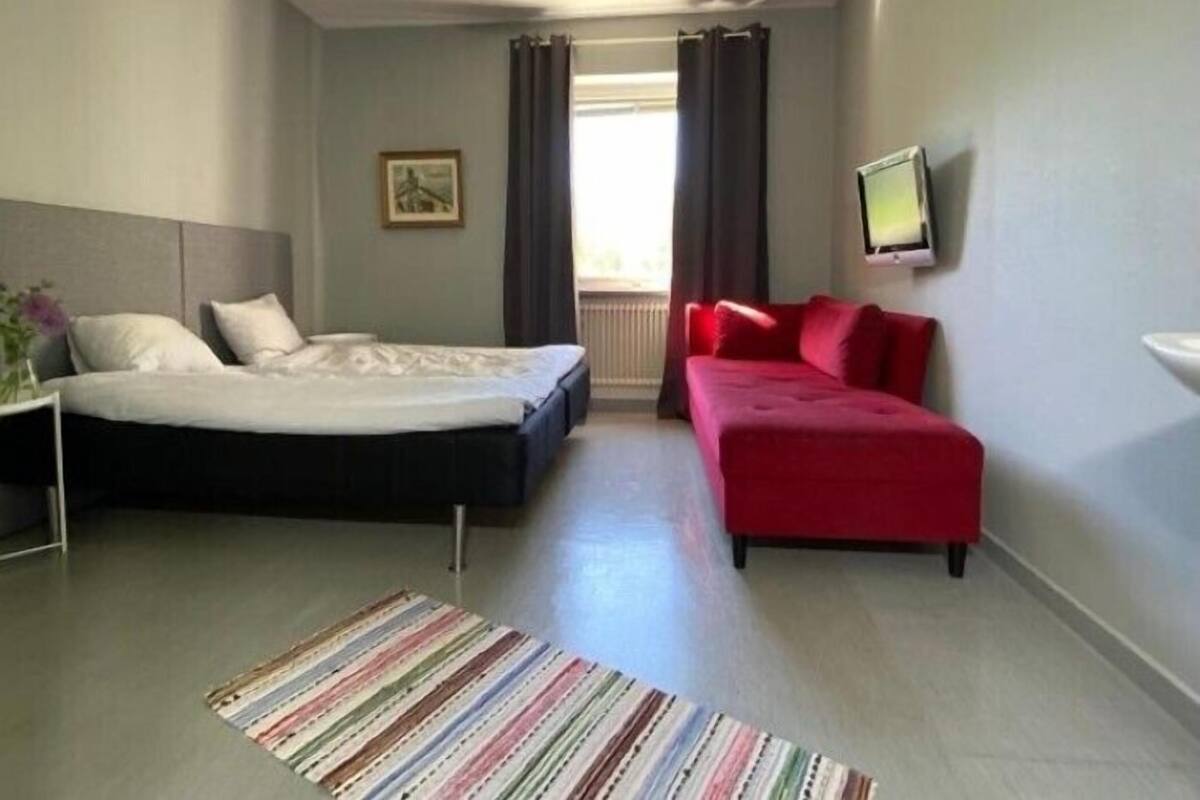 Ein Schlafzimmer mit einem Bett, einem roten Schlafsofa, einem Fernseher und einem Fenster mit Vorhängen.