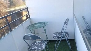 Een klein balkon met kunstgras, twee metalen stoelen met bloemenmotieven en een glazen tafel.