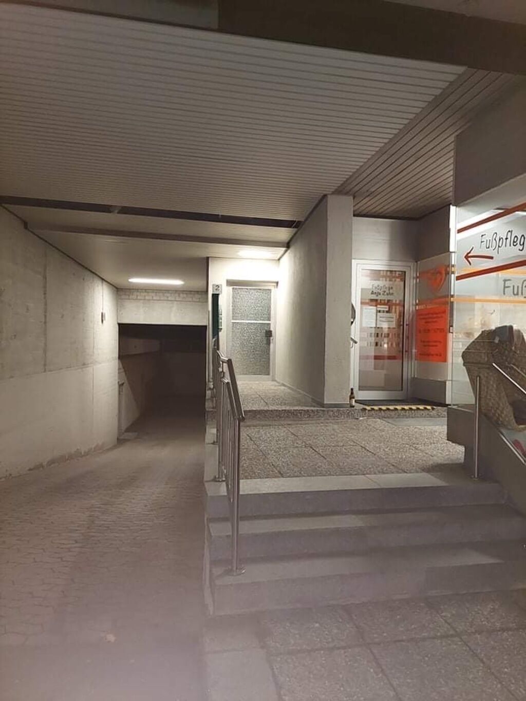 Um corredor subterrâneo com pouca luz, paredes de concreto, um guarda-corpo de metal e uma escada que leva a uma porta com o número 2.