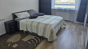Een slaapkamer met een bed, nachtkastje, een klein dressoir, een raam met gordijnen en uitzicht op gebouwen.
