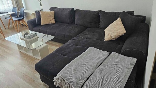 Een moderne woonkamer met een hoekbank, een glazen salontafel en twee fauteuils. Op de muur hangen abstracte kunstwerken en er staat een lamp in de hoek.