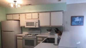 Una cocina compacta con electrodomésticos blancos, un microondas, una estufa y un refrigerador. Hay un lavamanos con grifo, un bloque de cuchillos y una pequeña silla blanca.