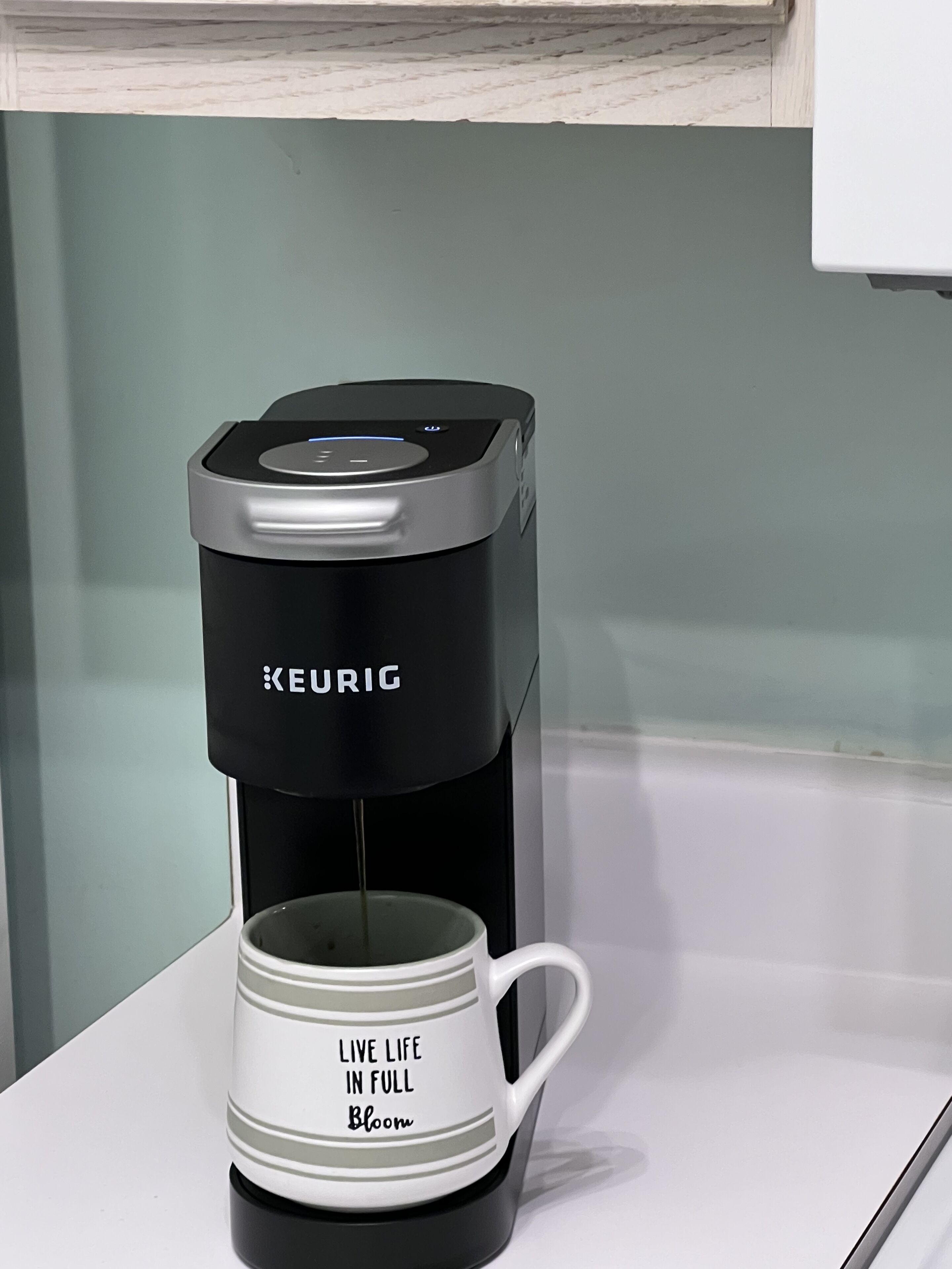 Una cafetera Keurig con una taza que dice 'Vive la vida en plena floración' sobre una encimera blanca.