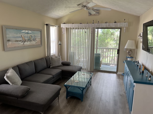A living room with a sectional sofa, a glass coffee table, a ceiling fan, and a view of the outdoors through a sliding glass door.