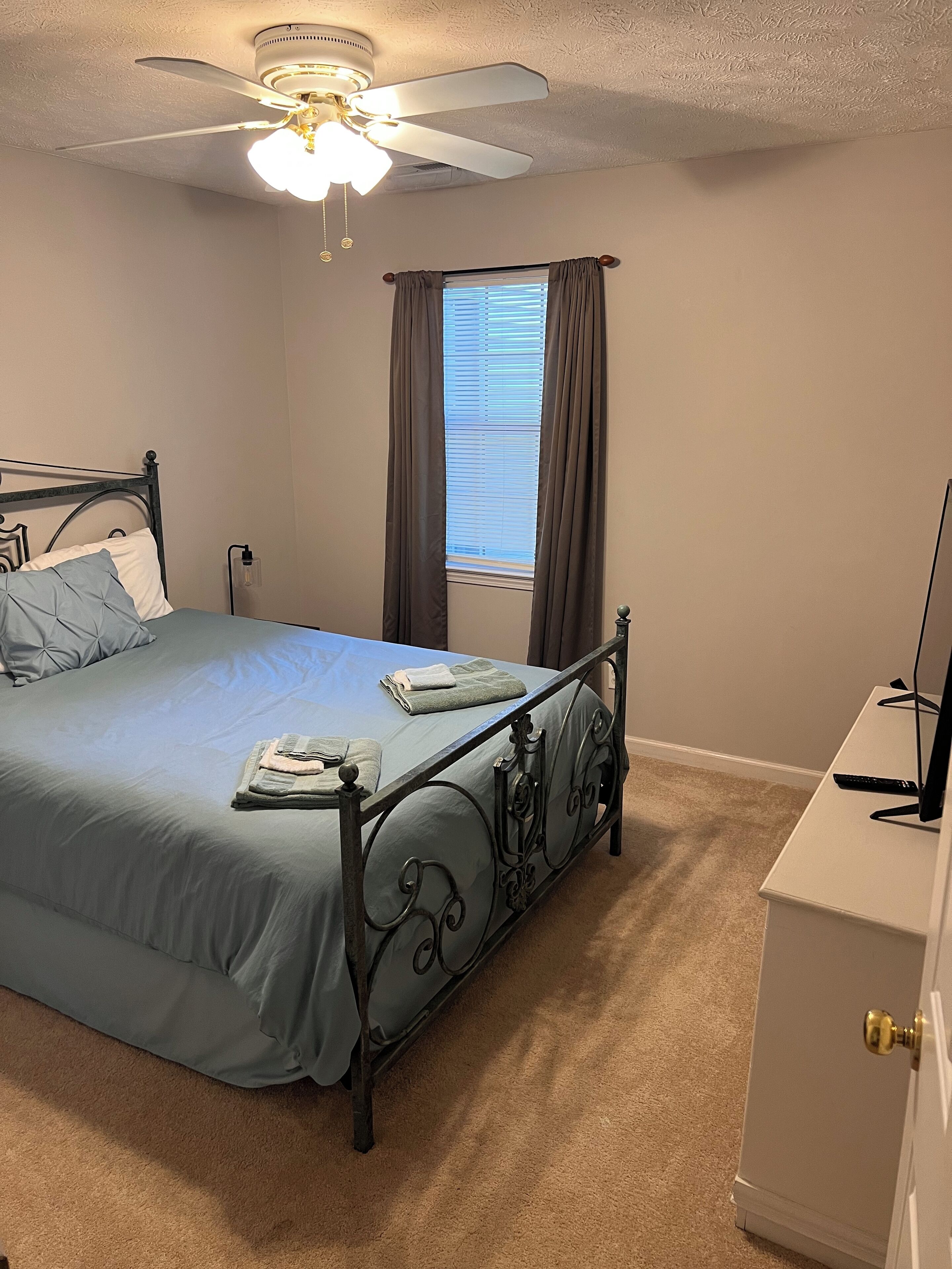 A bedroom with a bed, a ceiling fan, a window with curtains, a bedside table, and a TV.