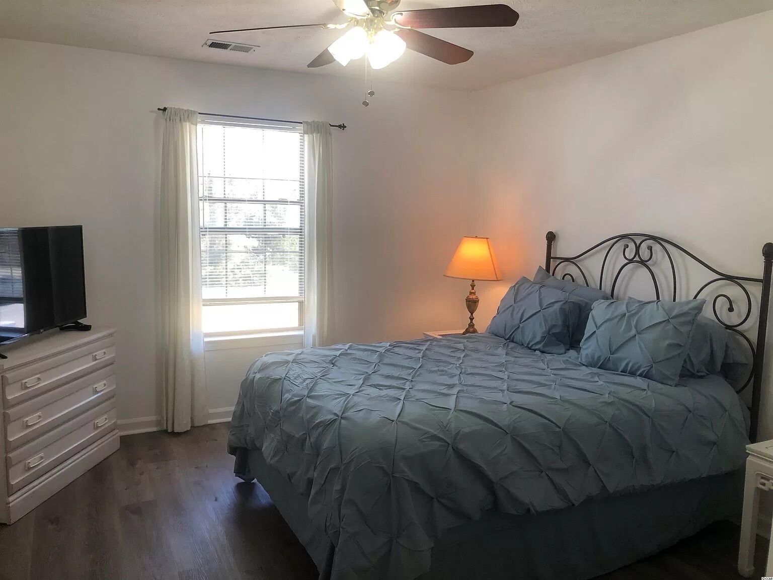A bedroom with a bed, a TV on a dresser, a window with curtains, a ceiling fan, and a bedside lamp.
