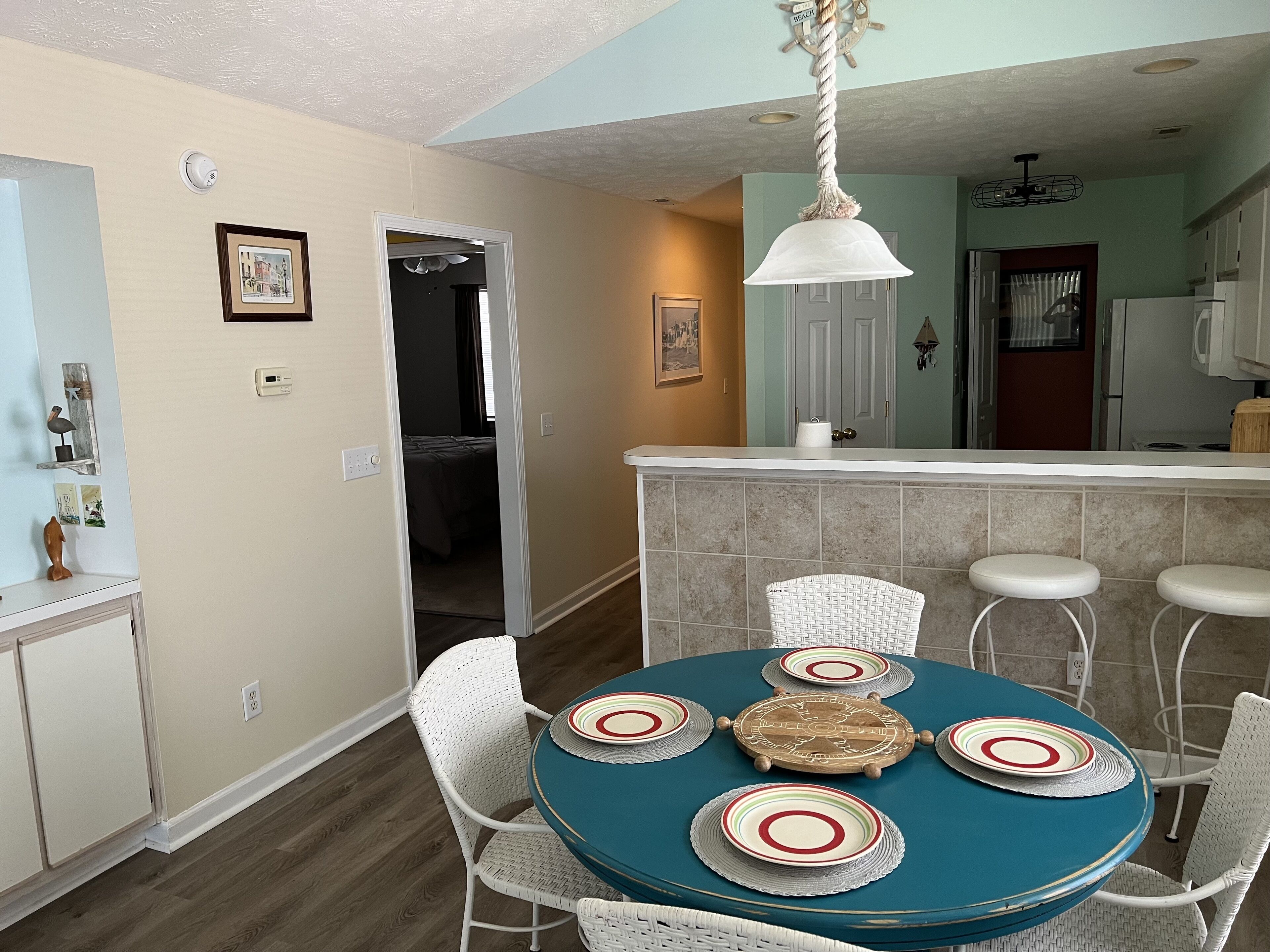 Uma área de refeições com uma mesa redonda, cadeiras de vime branco e toalha de mesa azul. Cozinha visível ao fundo, com bancada branca e luminária pendente.