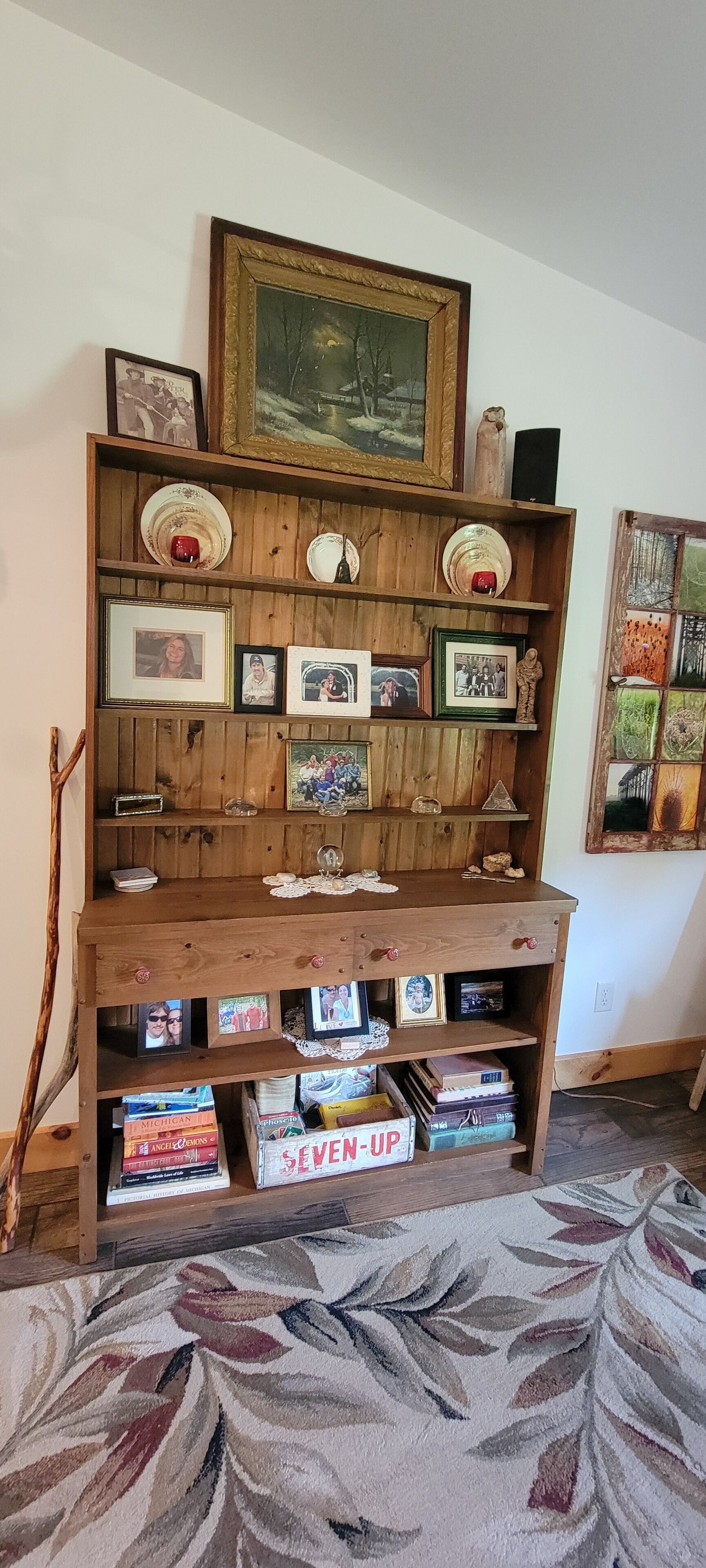 A wooden bookshelf with framed photos and books, a decorative plate, and a painting above it.