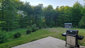 Un jardin avec un barbecue et un petit abri de jardin, le tout entouré d’arbres denses.
