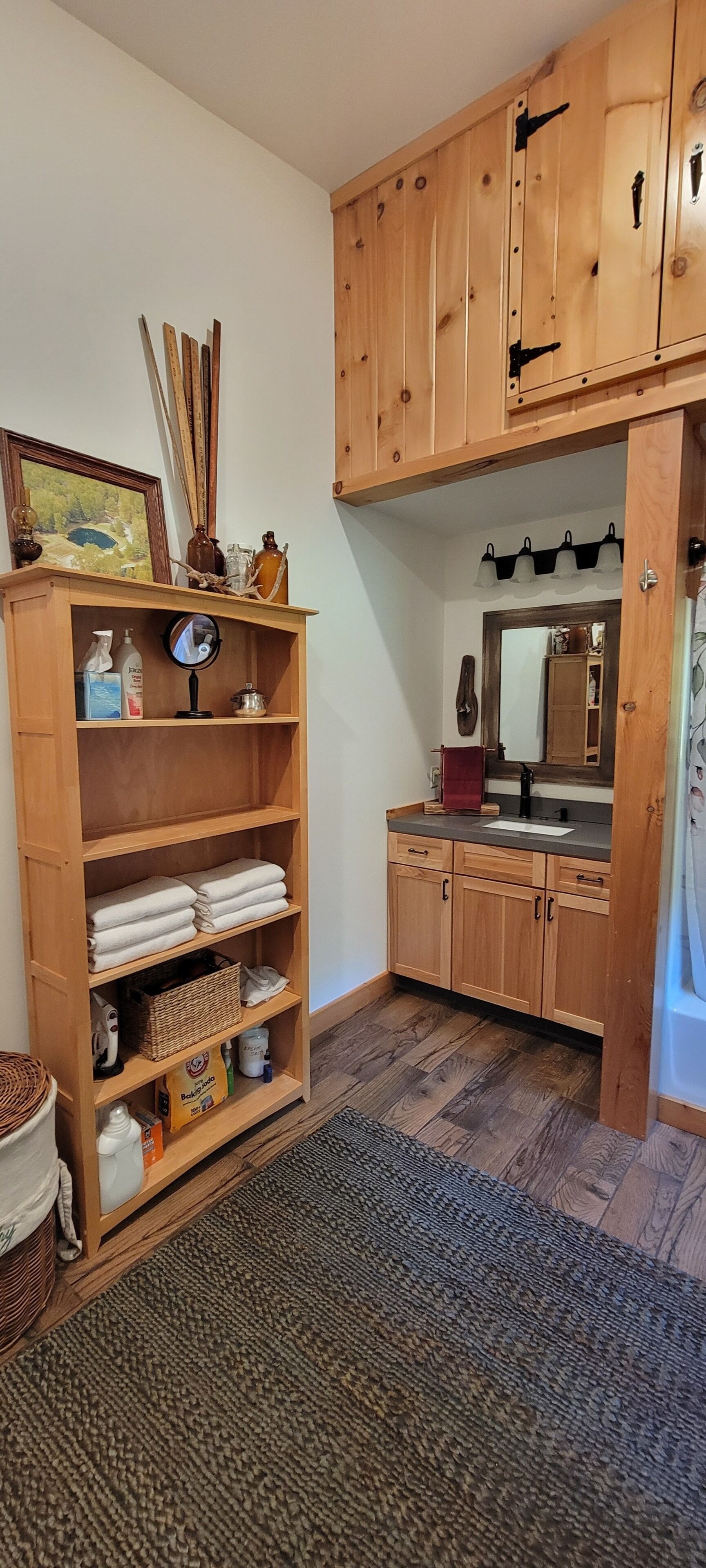 Een badkamer met houten kastjes, een wastafel en een spiegel. Er is een plank met handdoeken en toiletartikelen.