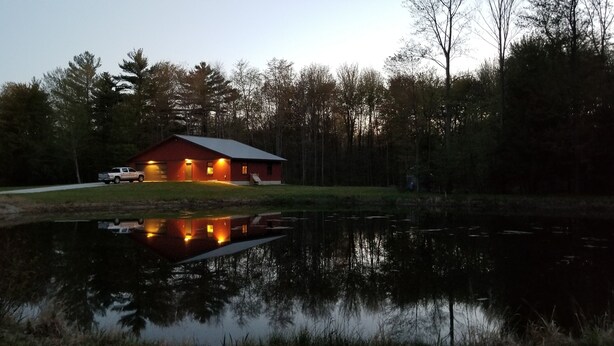 Une cabane rouge, avec un étang aux reflets argentés devant, entourée d’arbres.