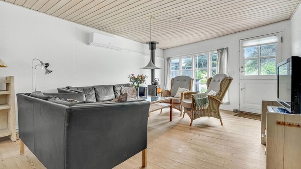 Ein modernes Wohnzimmer mit einem grauen Sofa, Korbstühlen, Holzboden und großen Fenstern.