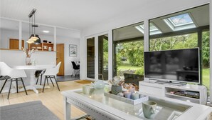 A modern living room with a large TV, a glass coffee table, and a dining area with a white table and chairs. - 2 bedroom cozy home in Dronningmølle (Dronningmølle)