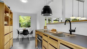 A modern kitchen with wooden cabinetry, a black pendant light, a sink, and a window offering a view of greenery. - 2 bedroom cozy home in Dronningmølle (Dronningmølle)