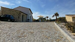 A stone-paved driveway leading to a white building with a tiled roof, a parked car, and a palm tree in the background.