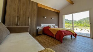 A bedroom with a bed, a sofa, a wardrobe, and a view of a green landscape through large windows.