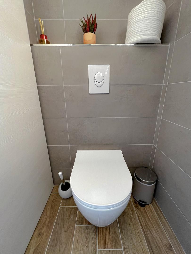 A compact bathroom with a toilet, a bidet, a trash bin, and a shelf with a plant and a bottle.