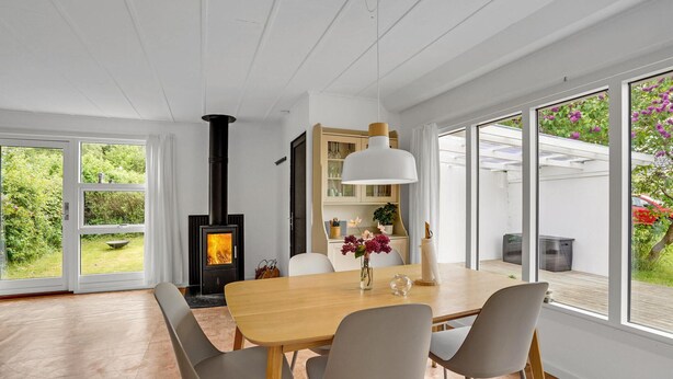 Uma sala de jantar moderna com uma grande mesa de madeira, cadeiras cinzas, uma luminária pendente branca e uma lareira a lenha preta.
