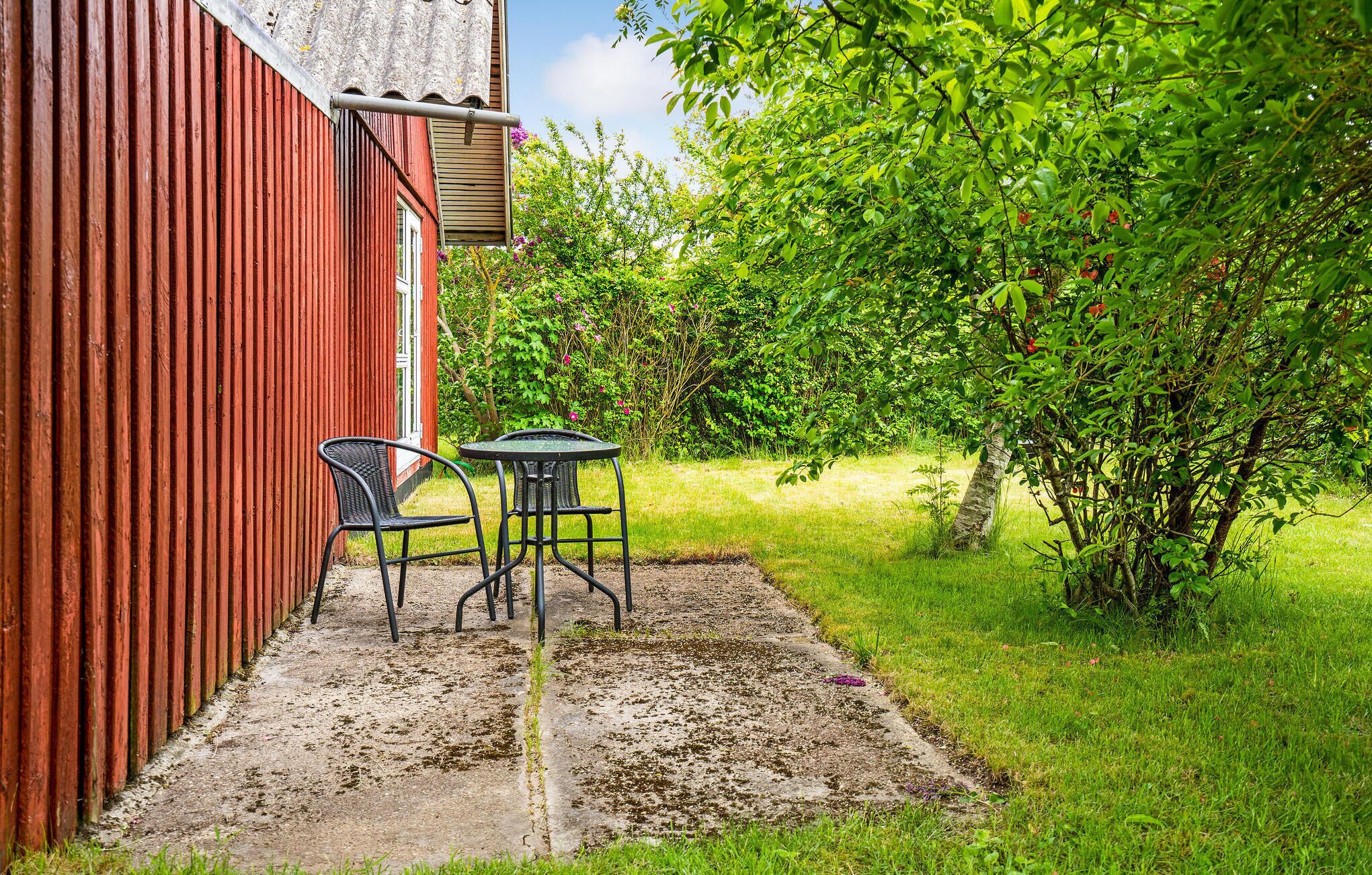 Een terras met twee stoelen en een tafel, een rood gebouw en weelderige begroeiing.