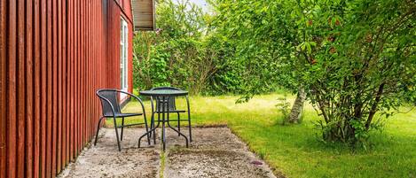 Een terras met twee stoelen en een tafel, een rood gebouw en weelderige begroeiing.