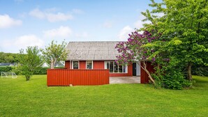 Uma casa de madeira vermelha com telhado cinza de telhas, um pequeno alpendre e um jardim com grama verde e flores roxas.