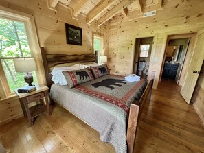 Quarto de cabana de madeira com cama, mesa de cabeceira, janela e porta que dá para um banheiro.