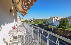 Un balcon avec une table et des chaises, donnant sur une rue bordée de voitures garées et d’immeubles.