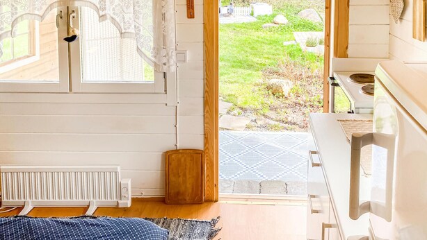A room with a wooden floor, a radiator, a window with lace curtains, a view of a garden, and a white stove.