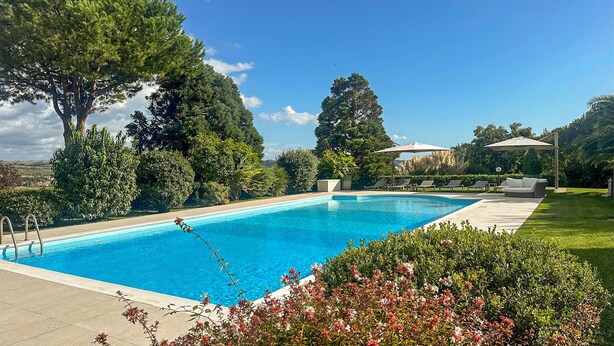 Una piscina rectangular con superficie de agua azul cristalina, rodeada por un césped cuidado y diversos árboles.